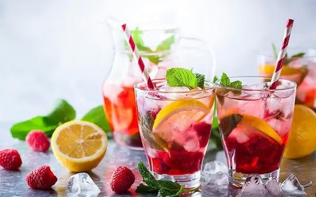 Two raspberry cocktails with jug in the background with fresh fruit garnishes
