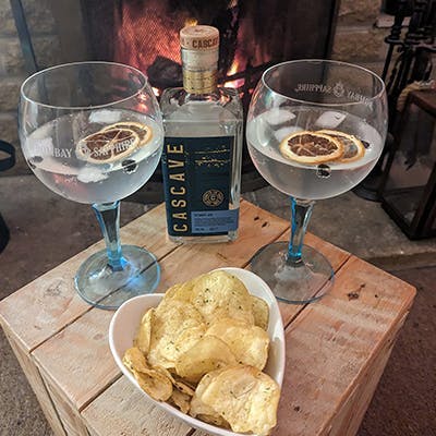 2 G&Ts and a bottle of cascave gin paired with a heart-shaped bowl of crisps in front of a roaring fire