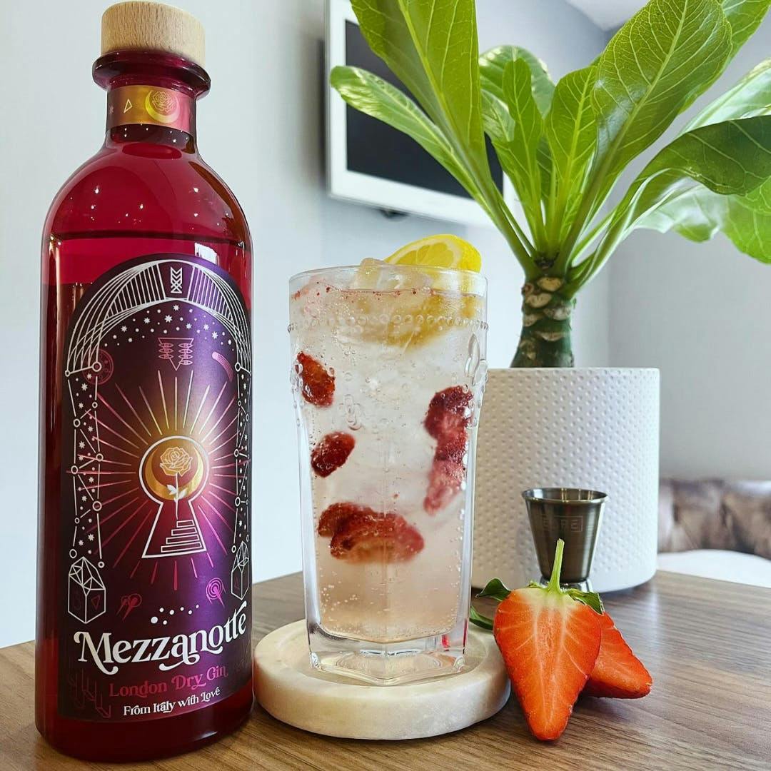 A bottle of February 2024's Gin of the Month with a G&T and strawberries in the foreground and a plant in the background