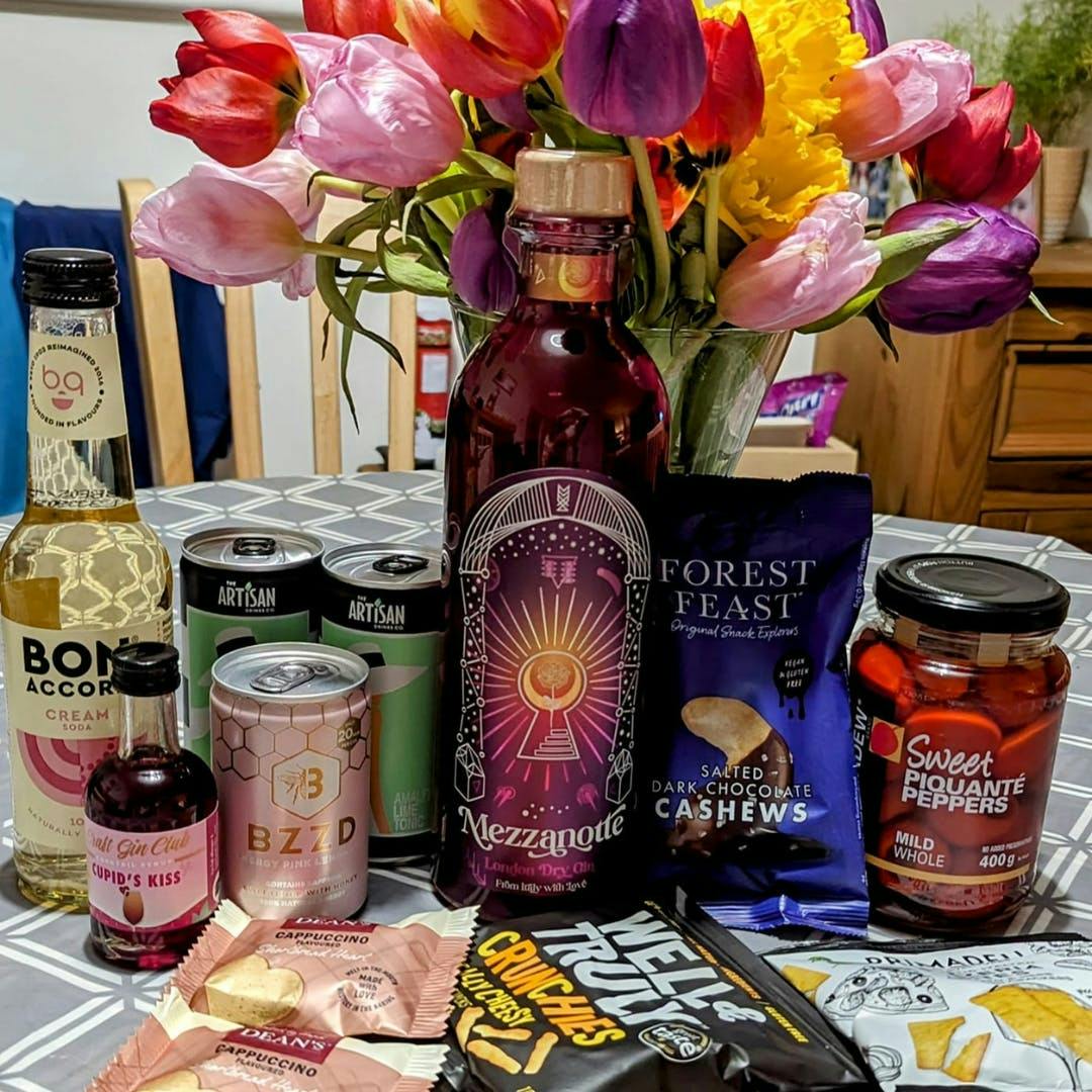 February's Gin of the Month box contents including a bottle of gin, snacks, tonics and mixers with a vase of tulips in the background.