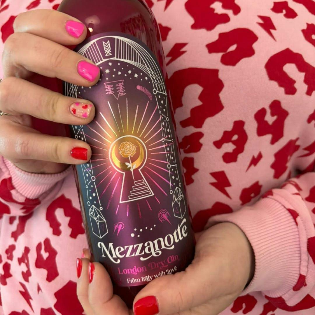 A bottle of Gin Mezzanotte (February 2024's Gin of the Month) held by someone in a pink animal print jumper and pink and red nails that match the pink bottle colour and jumper colour.