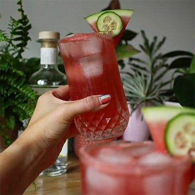 A delicious red coloured cocktail garnished with a slice of cucumber and watermelon