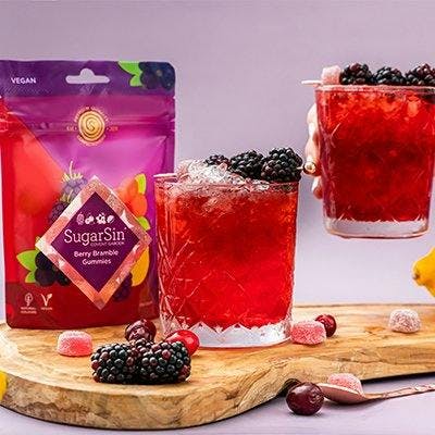 Two bramble cocktails with blackberries, next to a pouch of sugar sin berry bramble gummie sweets