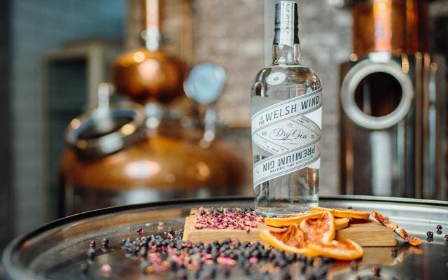 A bottle of In the Welsh Wind gin surrounded with botanicals with a copper still in the background.