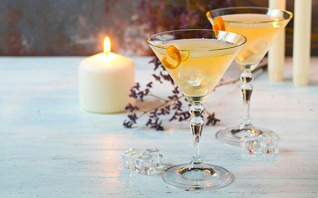 Two pale yellow cocktails in martini glasses with orange twist garnish