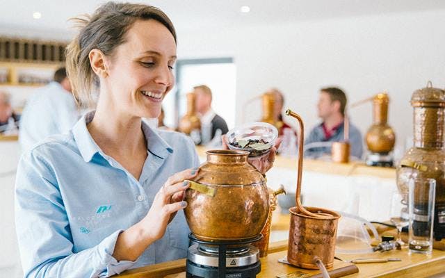 A smiling woman makes her own gin on a small still.
