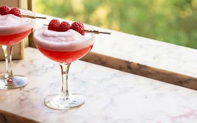 Pink foamy cocktail in a couple glass with a garnish of three speared fresh raspberries