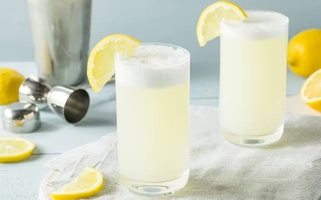 Two pastel yellow foamy cocktails in highball glasses with lemon wedge garnishes