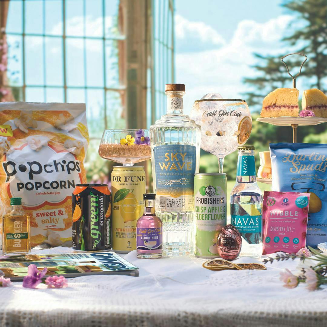A bottle of craft gin with a blue label and white horse design is surrounded by cocktails, cake and ingredients of a Gin of the Month box.
