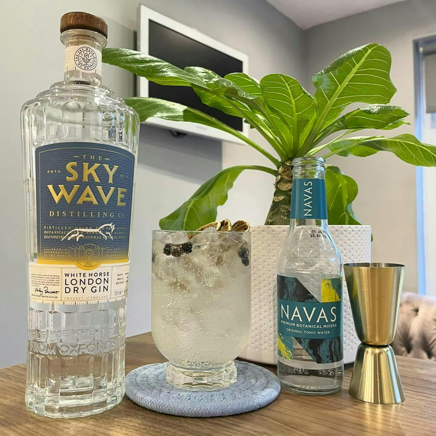 A bottle of Sky Wave Gin next to a gin and tonic and bottle of Navas tonic with an indoor plant in the background